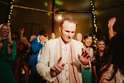 A man in a white jacket and scarf is dancing at a lively indoor event, surrounded by smiling people, with fairy lights strung around the venue, resembling the joyous atmosphere of a Kent wedding.