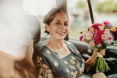 A smiling woman in a floral dress holds a bouquet of colorful flowers while seated in a car, capturing the joyous moment of her Kent wedding.