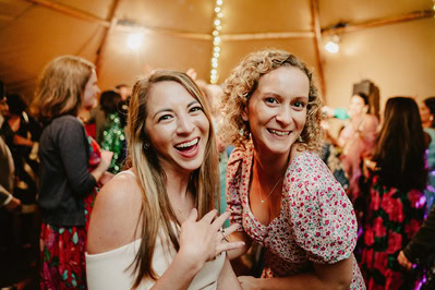 Two women smiling at a lively Kent wedding, surrounded by other guests in a festively lit space.