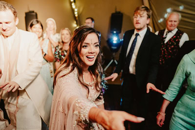 A woman in a festive outfit reaches out with a smile while people around her appear to be dancing and enjoying a Kent wedding.