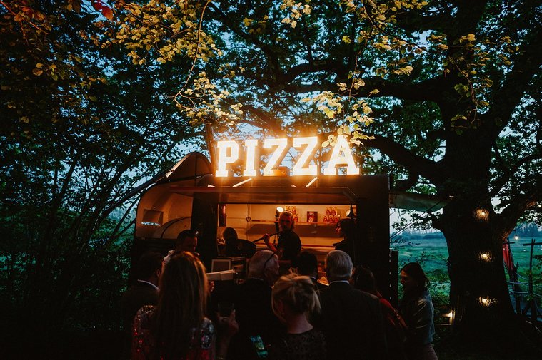 A crowd of people gather in front of a food truck at night, illuminated by a sign that reads "PIZZA" amidst trees and foliage, creating an enchanting atmosphere for those mindful of their wedding budgets.