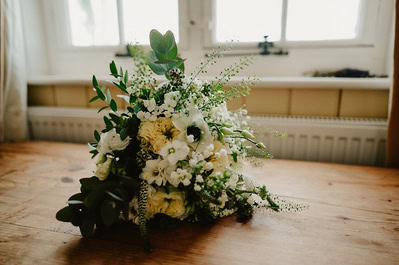 A bouquet of white and light green flowers with greenery is placed on a wooden table in front of a window, reminiscent of a Kent wedding at Beacon House.
