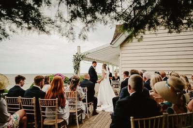 A bride and groom stand at an outdoor altar in front of seated guests during their Kent wedding ceremony near the beachside Beacon House.