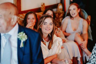 At a Kent wedding, a woman in white sits smiling among friends at an indoor event while a man in a navy suit with a flower boutonniere stands in the foreground.