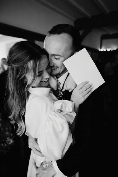 A couple in formal attire embrace closely, both smiling. One person holds a piece of paper, partially covering their face. The black-and-white photo captures a joyful moment at a Kent wedding, possibly at the elegant Beacon House.
