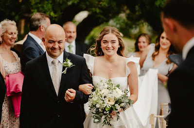 A bride and an older man, both smiling, walk down an outdoor aisle at a picturesque Kent wedding surrounded by well-dressed people. The bride holds a bouquet of white flowers as they make their way toward the charming Beacon House.