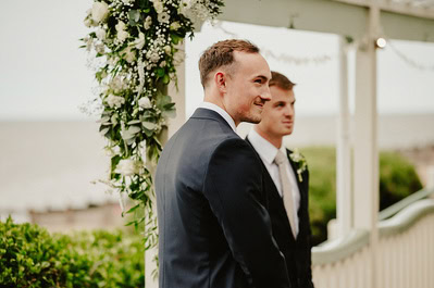 Two men in suits stand under a flower-adorned arbor overlooking a body of water, with greenery in the background. The serene ambiance hints at a beautiful Kent wedding, possibly at the romantic Beacon House location.