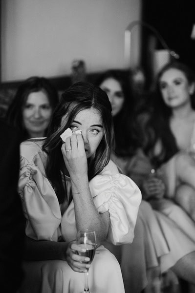 At a touching Kent wedding at Beacon House, a woman in a dress wipes her tears while holding a glass, with three other women seated behind her. The image is captured beautifully in black and white.