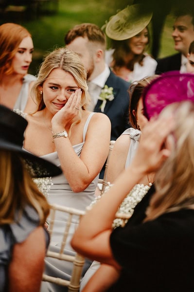 A woman in a light-colored dress and a gold watch wipes a tear from her eye at the Kent wedding, held at the picturesque Beacon House, surrounded by other attendees dressed in formal attire.