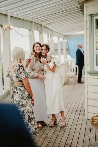 Two women, one in a white dress and the other in a light gray outfit, smile and pose on the wooden porch adorned with string lights at Beacon House during a Kent wedding. In the background, another person in a floral dress stands beside a man in a suit.
