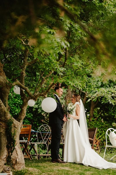 A bride and groom stand facing each other under a tree with white lanterns, holding a bouquet, during their enchanting outdoor Kent wedding at Beacon House.