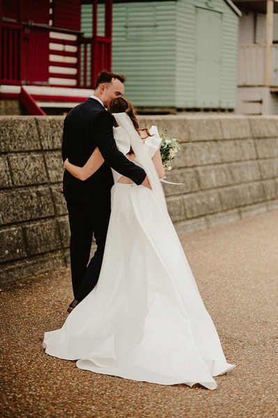 A bride in a white dress and groom in a black suit embrace while walking on a paved path at Beacon House, with colorful buildings in the background, during their picturesque Kent wedding.