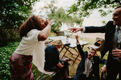 At a Kent wedding by Beacon House, a woman drinks from a glass while another woman sits looking down. Two men—one seated on the ground and one standing—react animatedly outdoors, with a serene lake and lush trees in the background.