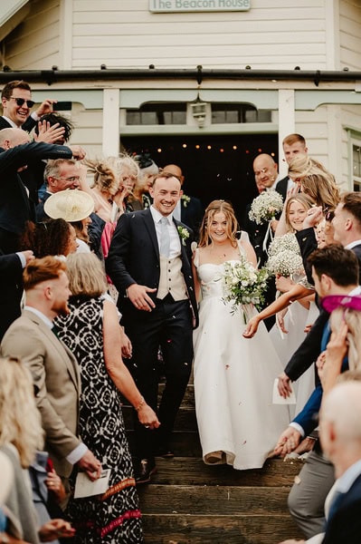 A newly married couple, dressed in a suit and wedding dress, smiles and walks down the steps of Beacon House, surrounded by joyous family and friends celebrating their Kent wedding.