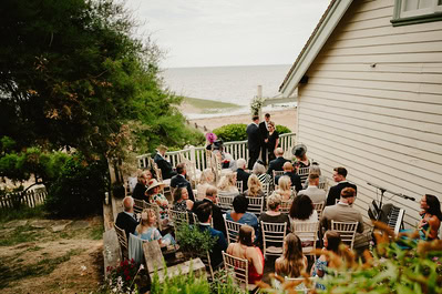 Outdoor Kent wedding ceremony by the sea, with guests seated on white chairs facing a couple standing next to an officiant near Beacon House.