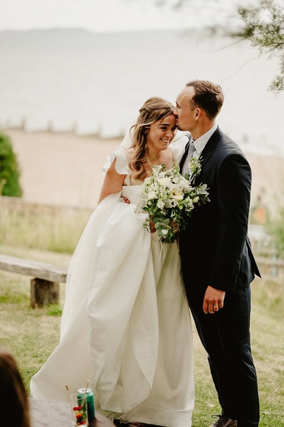 A couple in wedding attire stands close together outdoors at Beacon House, with the groom kissing the bride's forehead. The bride holds a bouquet of flowers, and a beach along with lush greenery can be seen in the background of their beautiful Kent wedding.