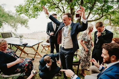 At a beautiful Kent wedding at Beacon House, a group of people in formal attire gather outdoors. One man stands and raises his arms in celebration, while others around him smile and interact joyfully.