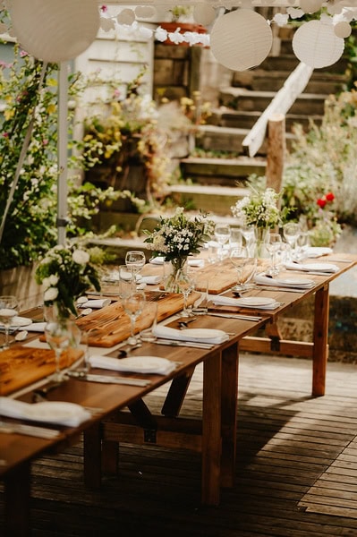 A wooden outdoor table is set with plates, glasses, cutlery, and floral centerpieces under hanging white paper lanterns, reminiscent of a charming Kent wedding at Beacon House, with a staircase in the background.