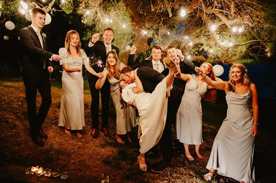 A newlywed couple shares a kiss while others in the wedding party stand around them, some holding sparklers under the tree covered in string lights at night, capturing the magic of their Kent wedding at Beacon House.