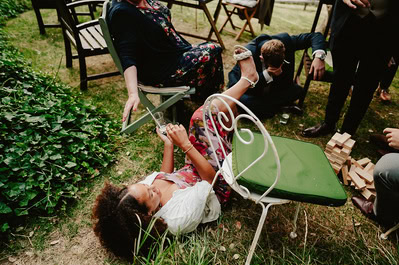 At a Kent wedding at Beacon House, a person lies on the ground next to a fallen chair, laughing and holding a drink, while others around them, dressed formally, react.