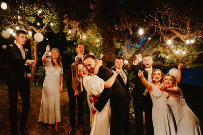 A wedding party poses under string lights at night at Beacon House. The bride and groom embrace at the center while the bridesmaids and groomsmen celebrate around them with joyful expressions, capturing a beautiful Kent wedding.