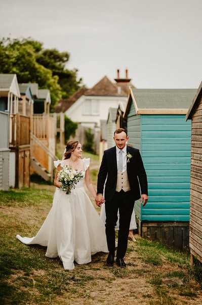 A bride in a white dress and a groom in a black suit and beige vest walk hand in hand past wooden beach huts at Beacon House, celebrating their Kent wedding.