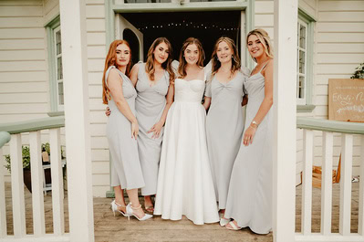 Five women in formal dresses stand together in front of Beacon House with its white façade and green trim. Four wear light gray dresses, and one in the middle wears a white gown, perfectly capturing the elegance of a Kent wedding.