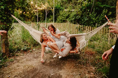 Three women in dresses laugh and try to balance on a hammock in a garden setting, surrounded by lush greenery and twinkling string lights, enjoying the whimsical charm of a Kent wedding at Beacon House.