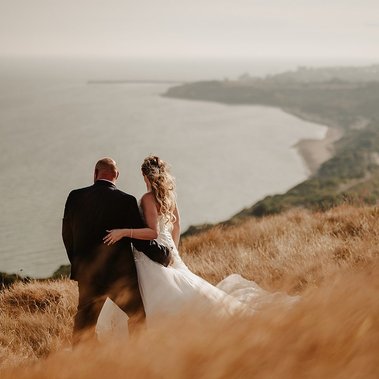 A bride and groom stand closely together on a grassy hilltop overlooking a coastal landscape with the ocean and distant shoreline visible. The scene, captured by a wedding photographer, is serene and taken during sunset.