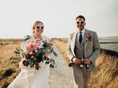 A bride and groom wearing heart-shaped sunglasses walk outdoors on a sunny day. The bride holds a large bouquet of flowers, and both are smiling as their wedding photographer captures the joyful moment.