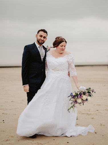 A wedding photographer captures a bride in a white dress holding a bouquet, standing next to a groom in a dark suit on a beach with a cloudy sky.