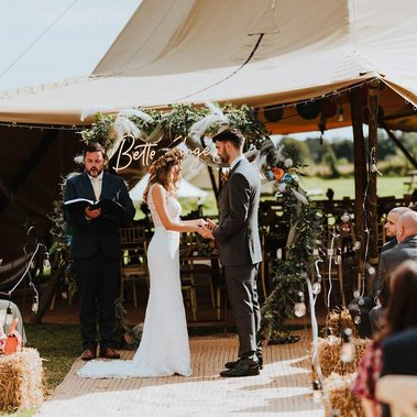 A bride and groom stand facing each other holding hands at an outdoor wedding ceremony. An officiant stands beside them, and a photographer captures the moment against a canopy adorned with foliage and decor in the background.