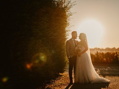 A photographer captures a bride and groom standing close together, silhouetted by the setting sun next to a tall hedge. The bride holds a bouquet and wears a flowing gown with a veil, adding to the magic of their wedding day.