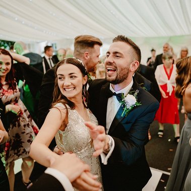A bride and groom, smiling and holding hands, dance together at their wedding reception in a tent. Captured by the wedding photographer, guests in the background are also dancing and celebrating.