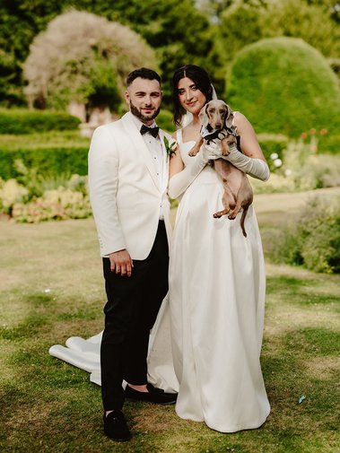 A couple in formal attire stands outdoors on a lawn, the man wearing a white suit and black pants, the woman in a white dress holding a small brown dog, as their wedding photographer captures the moment.