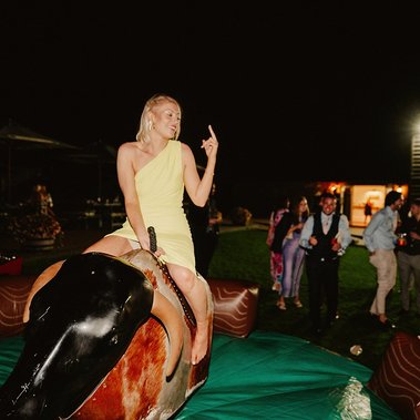 A woman in a yellow dress rides a mechanical bull at night while holding up one finger, all captured perfectly by a skilled photographer. People in the background are watching and smiling.