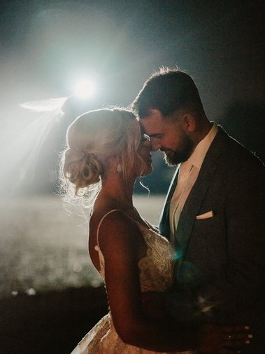 A bride and groom stand close, touching foreheads, illuminated by a bright light in the dark. Captured by their photographer, the bride wears a white dress with an updo hairstyle, and the groom is in a grey suit with a white shirt, making this wedding moment unforgettable.