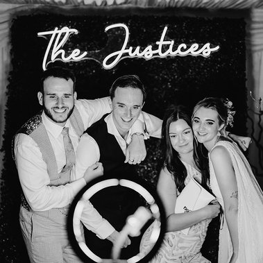 Black and white photo of four people posing in front of a sign that reads "The Justices." A ring light, often used by photographers, is visible at the bottom of the image.
