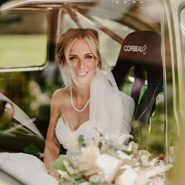 A bride in a white wedding dress and veil sits in the driver's seat of a vehicle, smiling at the camera. She holds a bouquet of flowers on her lap, perfectly framed by the wedding photographer's keen eye.