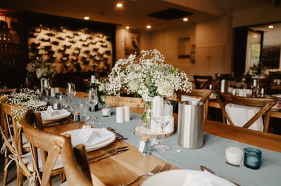 A decorated dining table with white flowers, candles, silverware, and glasses set in a dimly lit restaurant at the elegant Chapel House Estate, with a wine display on the wall in the background—perfect for an intimate wedding celebration.