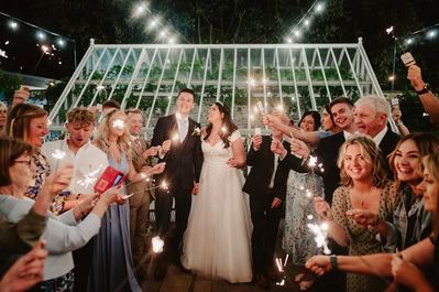 A newlywed couple stands at the center, surrounded by people holding sparklers under string lights and a glass structure at the charming Chapel House Estate, during an enchanting evening wedding celebration.