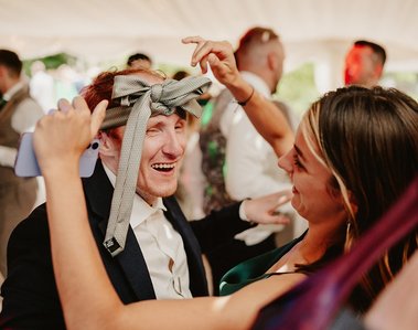 Two people at a wedding party; the man has a tie wrapped around his head and is smiling, while the woman holding a smartphone smiles back at him. People, including the wedding photographer, are visible in the background.