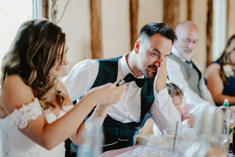 A bride shows something on her phone to the groom, who appears emotional, during their wedding reception. Other guests are seated at the table, as a wedding photographer captures this intimate moment.