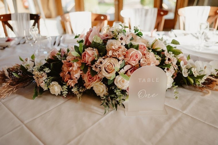 A floral centerpiece with roses and greenery adorns a table with a "Table One" sign, perfect for the wedding photographer to capture.