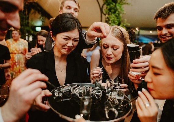 Group of people in semi-formal attire sampling drinks around a tray, with varied reactions, at what appears to be a wedding, possibly captured by a wedding photographer.