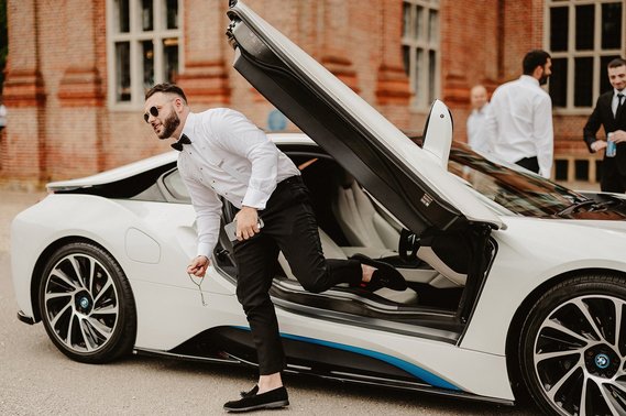 A man in formal attire exits a white sports car with upward opening doors, likely heading to a wedding. Other men in dress shirts are visible in the background near a brick building.