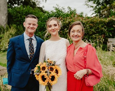 Three people are posing for a photo outside, likely taken by a wedding photographer. The person in the center, wearing a white dress and holding a bouquet of sunflowers, stands between two others: one in a blue suit on the left and another in a red outfit on the right.