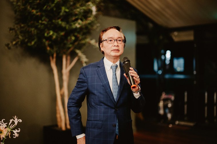 A man in a blue checkered suit and tie delivers a wedding speech into a microphone indoors, with a potted plant and dim lighting providing an intimate backdrop.