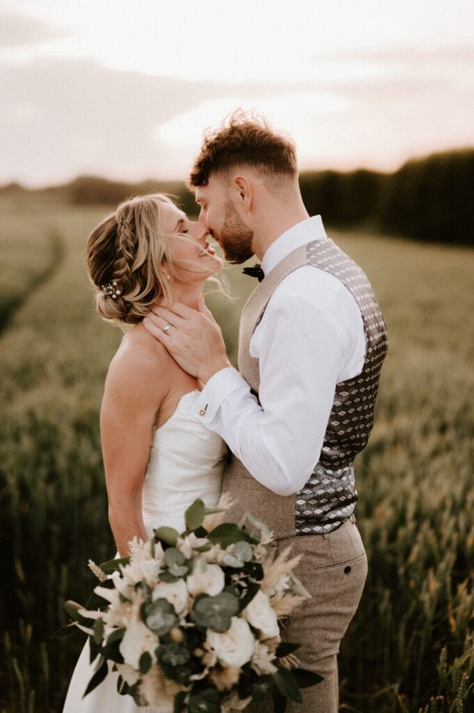 A bride and groom embrace in a sunlit field, captured perfectly by a Kent wedding photographer. The bride holds a bouquet of white and green flowers as the groom, clad in a vest and bow tie, gazes into her eyes. They are enveloped by tall grass and the soft hues of a sunset sky. Image by Pearce Wedding Photography.