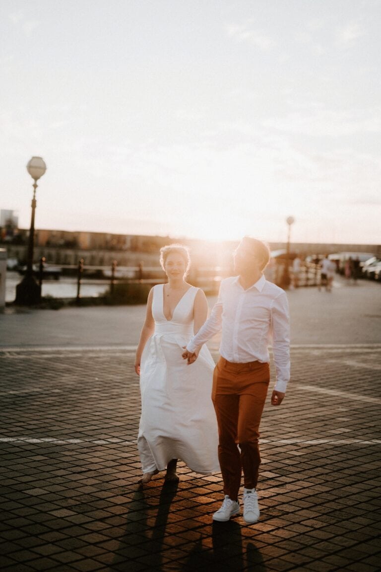 A couple walks hand in hand on a sunlit paved walkway, with the woman wearing a white dress and the man in a white shirt and brown pants.
